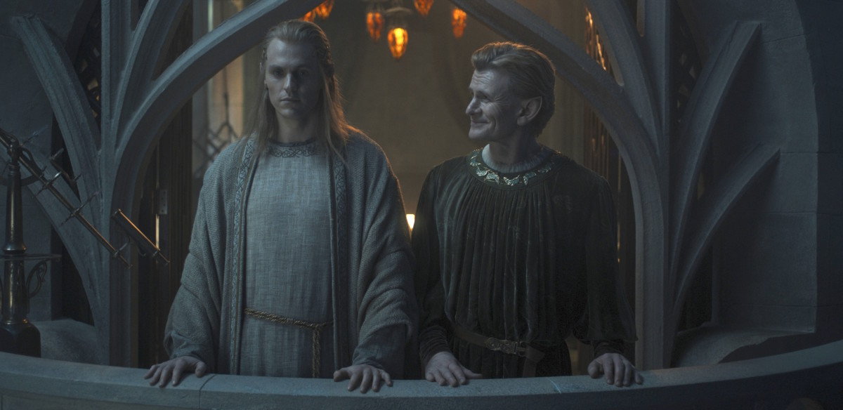 Charlie Vickers and Charles Edwards as Sauron and Celebrimbor in Rings of Power Season 2, standing together on a balcony. Celebrimbor looks at Sauron happily, while Sauron refuses to look at him