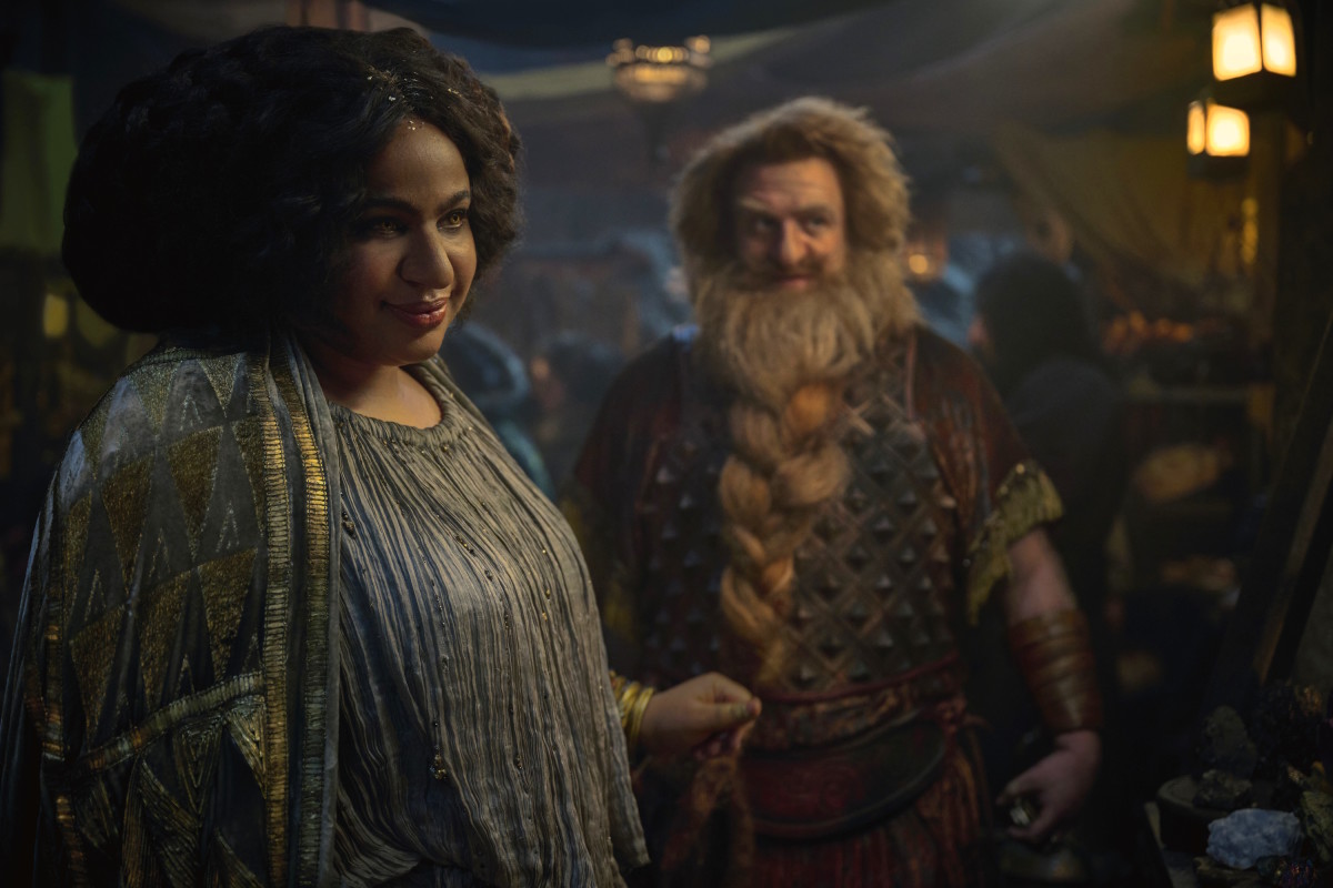 Sophia Nomvete and Owain Arthur as Disa and Prince Durin in Rings of Power Season 2, haggling with a market stall owner in a busy Khazad-dum street