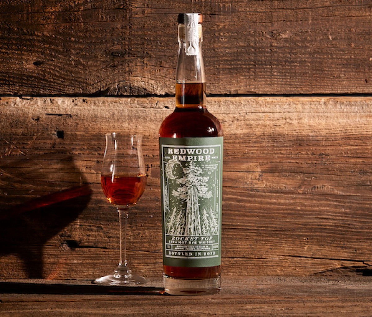 Bottle of Redwood Empire Rocket Top Bottled-in-Bond Rye with filled stem glass in front of wood slat background