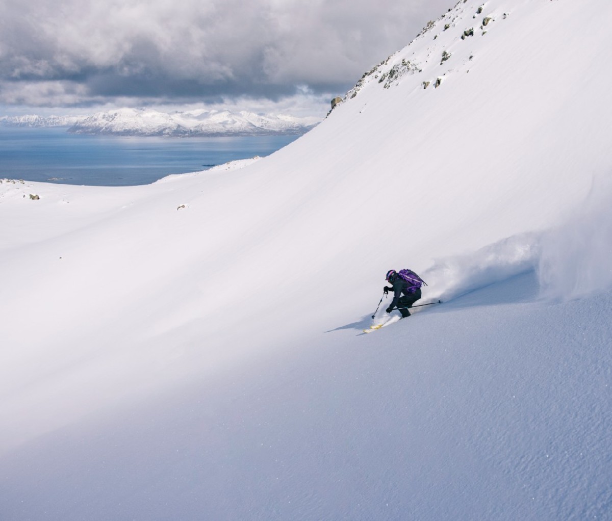 Skier heads down a slope of powder in Northern Norway with coastal waters in the far background.