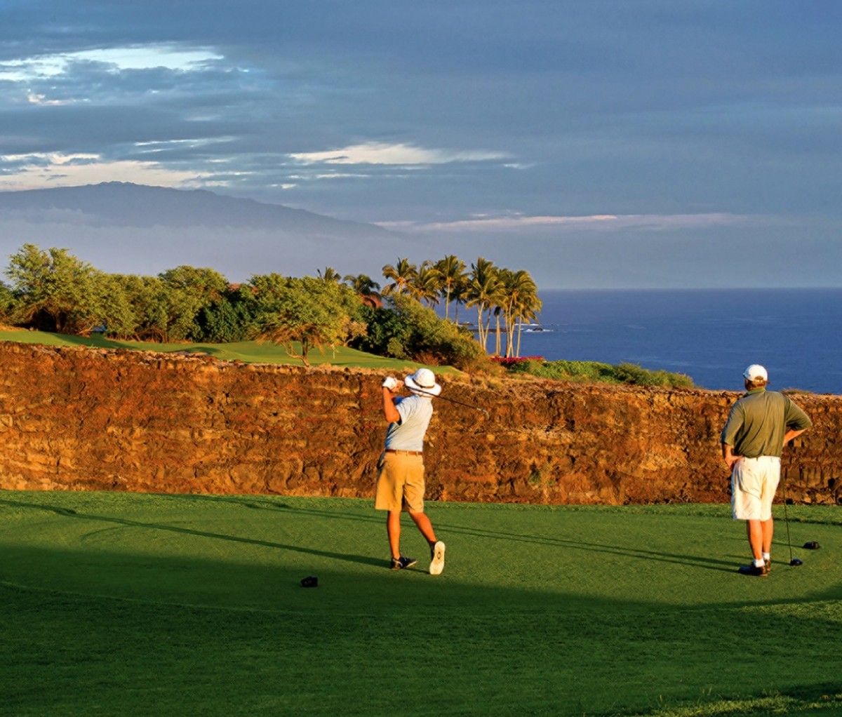 Two golfers, one taking a swing, at a golf course tee box overlooking a cliff. Golfing in Hawaii