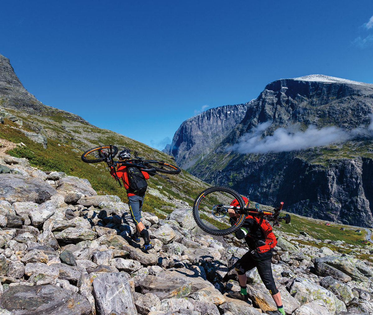 Two mountain bikers in red jackets carrying bikes on shoulders up steep mountainside