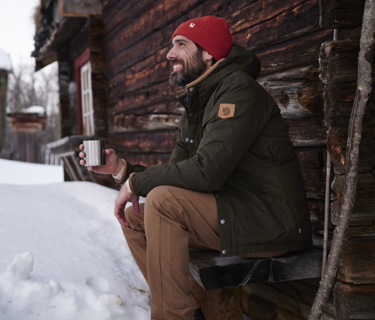 Man wearing red beanie, green waxed jacket, and tan trousers outside cabin in snow