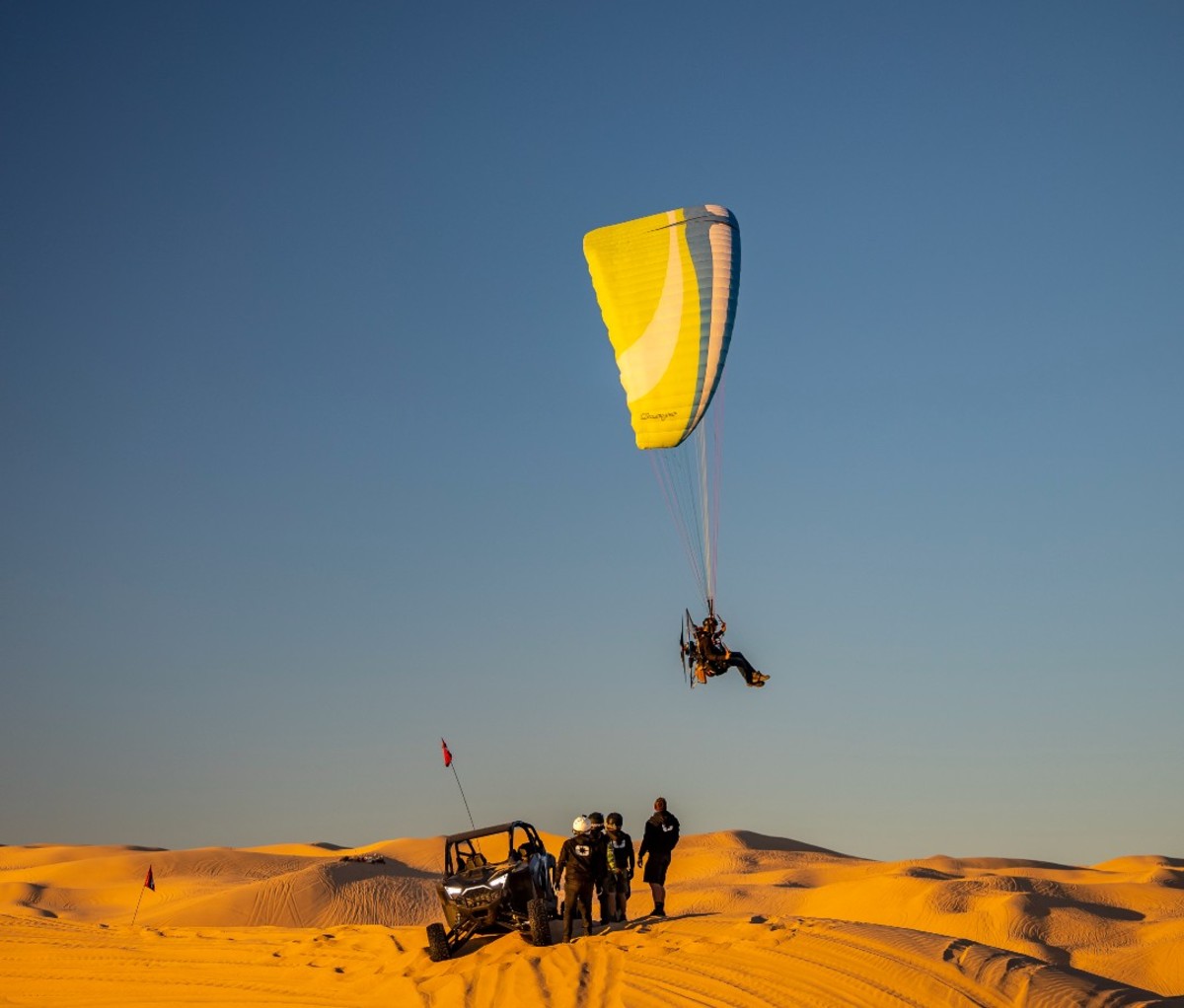 Camp RZR attendants in the dunes, standing beside vehicle and parasailing overhead.