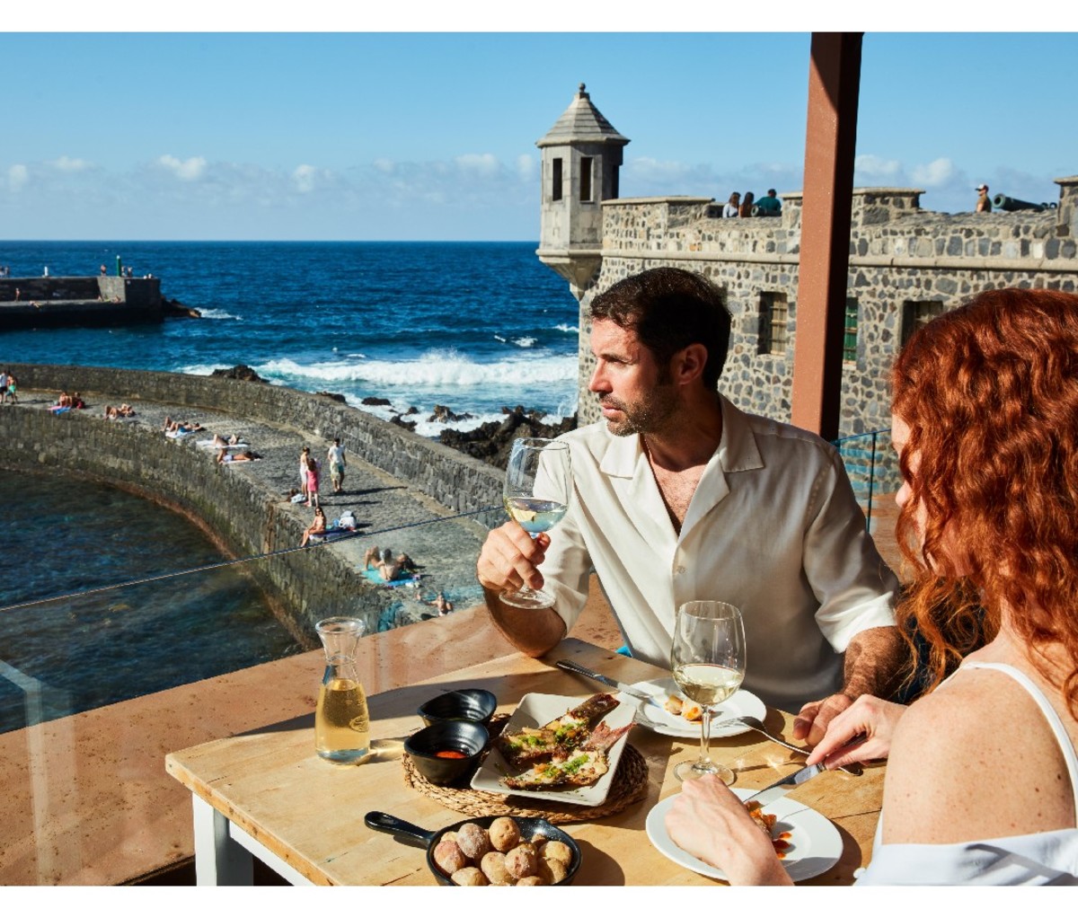 Couple dining on the coast of Tenerife, with castellated structure in background.