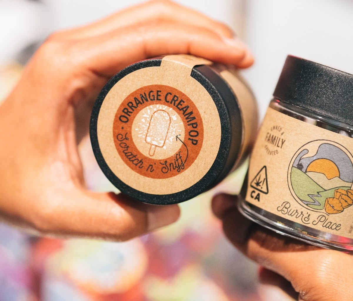 Hand holding a container of cannabis labeled with a scratch and sniff sticker.