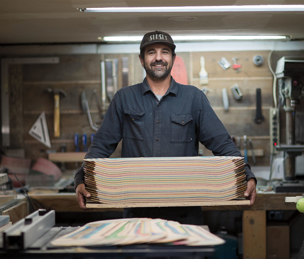 Young caucasian man wearing baseball hat and blue button-down smiling holding stacks of skateboard boards