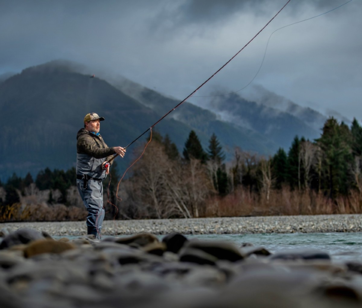 Man in waders fly fishing in a river with mountains and storm clouds in the background.