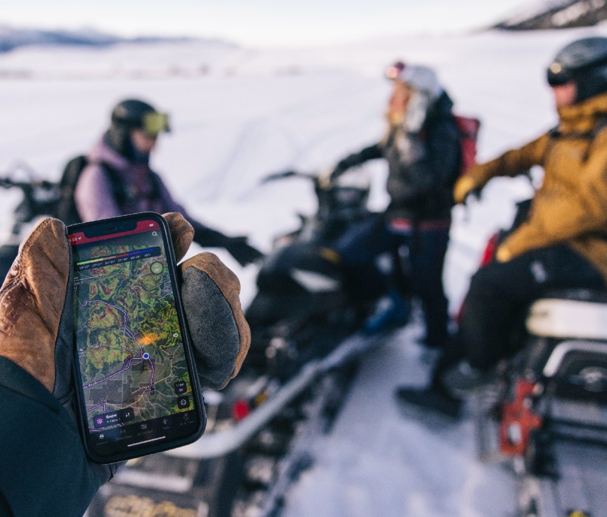 Hand displays navigation app on a phone with snowmobilers in the background.