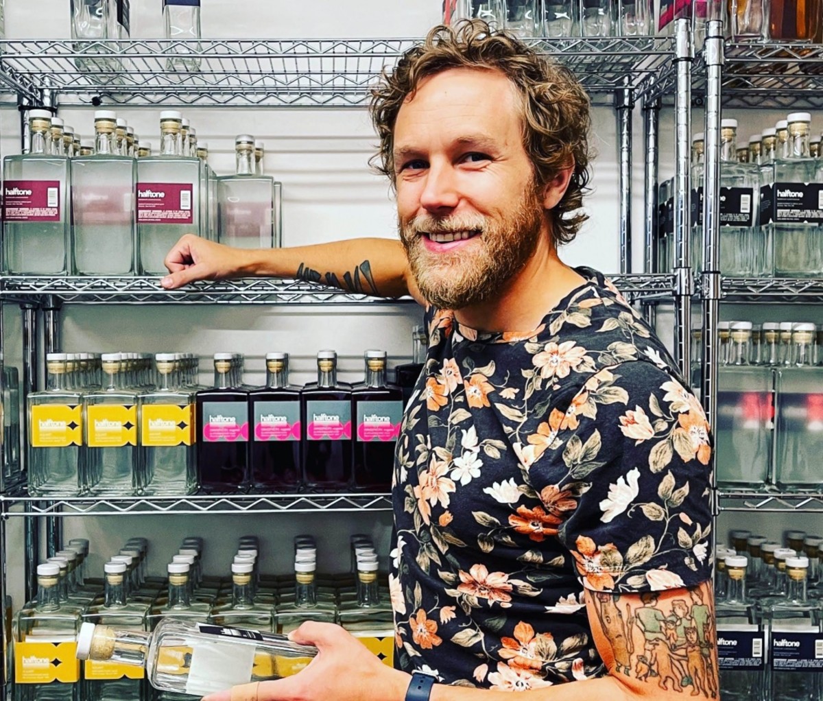 Bearded man smilling while holding a clear bottle of aquavit liquor in front of shelving holding more liquor bottles.
