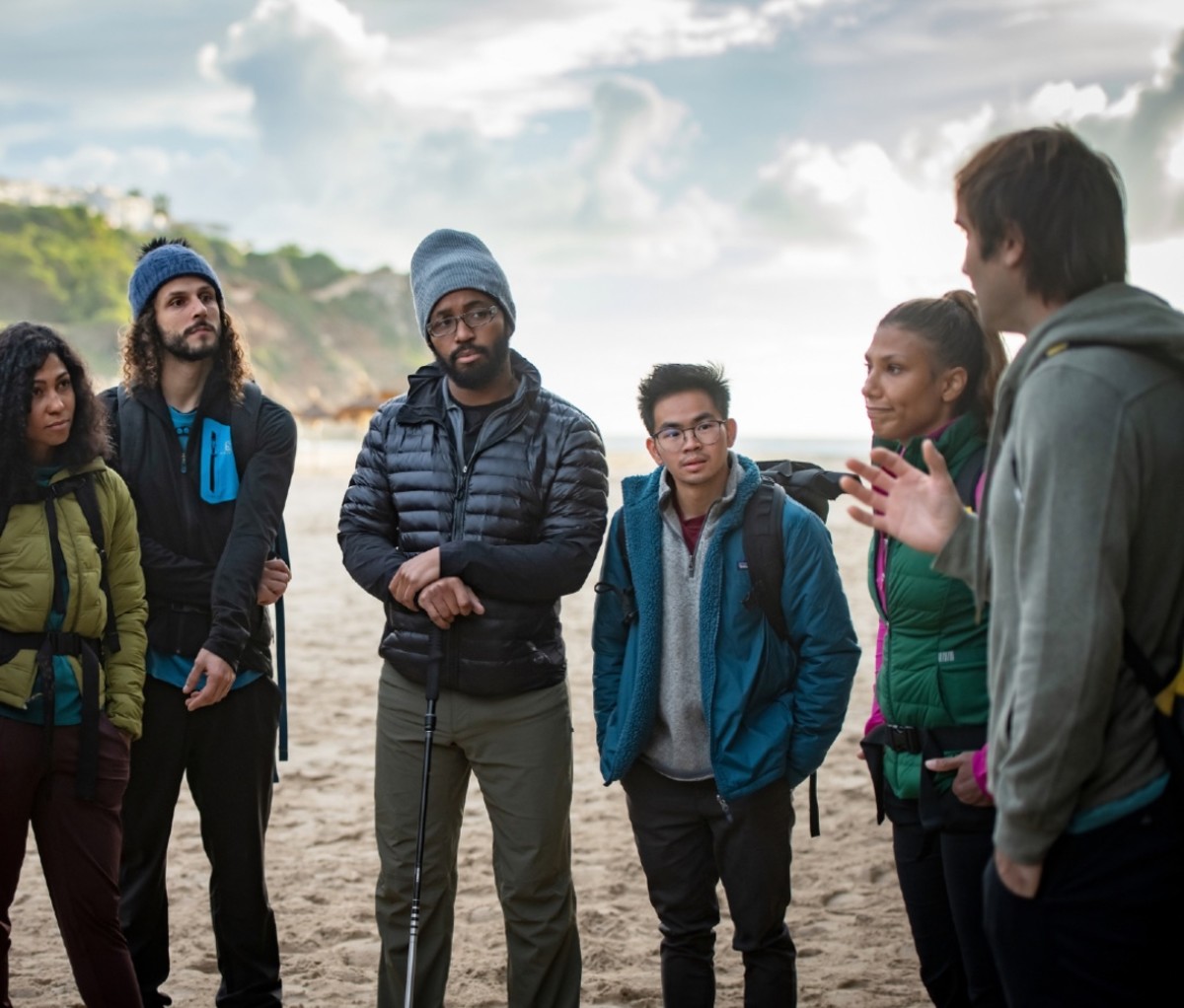 A group of rock climbers in jackets and hats stand around while another climber instructs them.