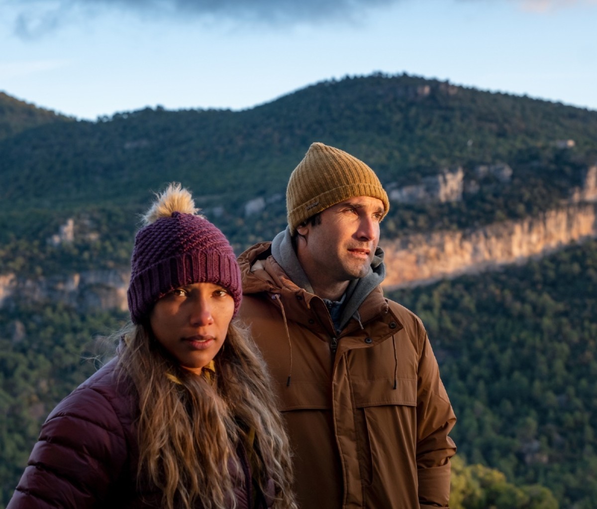 Man and a woman with hats and jackets stand in the mountains with a forest ridge line in the background.