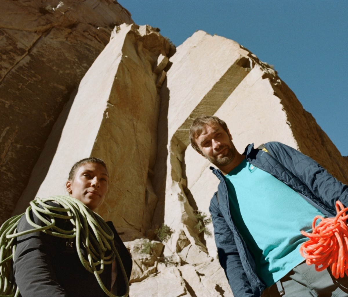 A female and male rock climber stand at the bottom of a steep ascent with ropes in their hands.