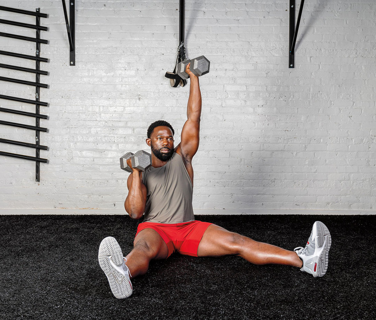 Black man wearing gray tank and red shorts doing Seated Straight-leg Bilateral Shoulder Press