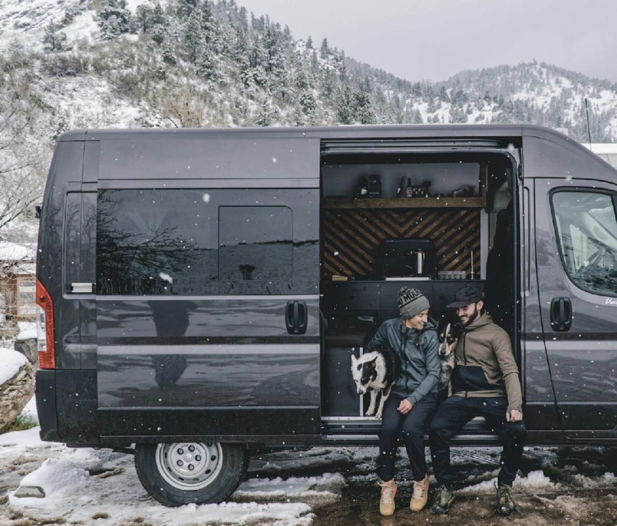 Couple sitting a doorway of a camper van with snowy mountains in the background.