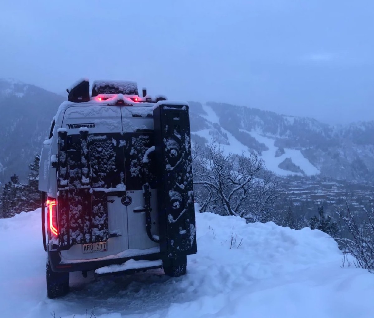 Snow-covered camper van with ski slopes in the background.